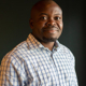 A Black man smiling in front of a dark grey background