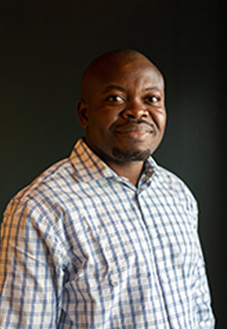 A Black man smiling in front of a dark grey background
