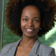 A Black woman with long hair wearing a gray blazer in front of a green background
