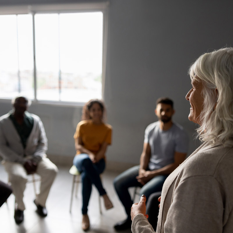 resources-mobile A grey haired woman talking in a group therapy setting