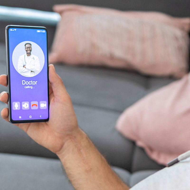 telehealth-mobile A hand holding a phone with a picture of a man in a lab coat that says 'doctor' on the screen